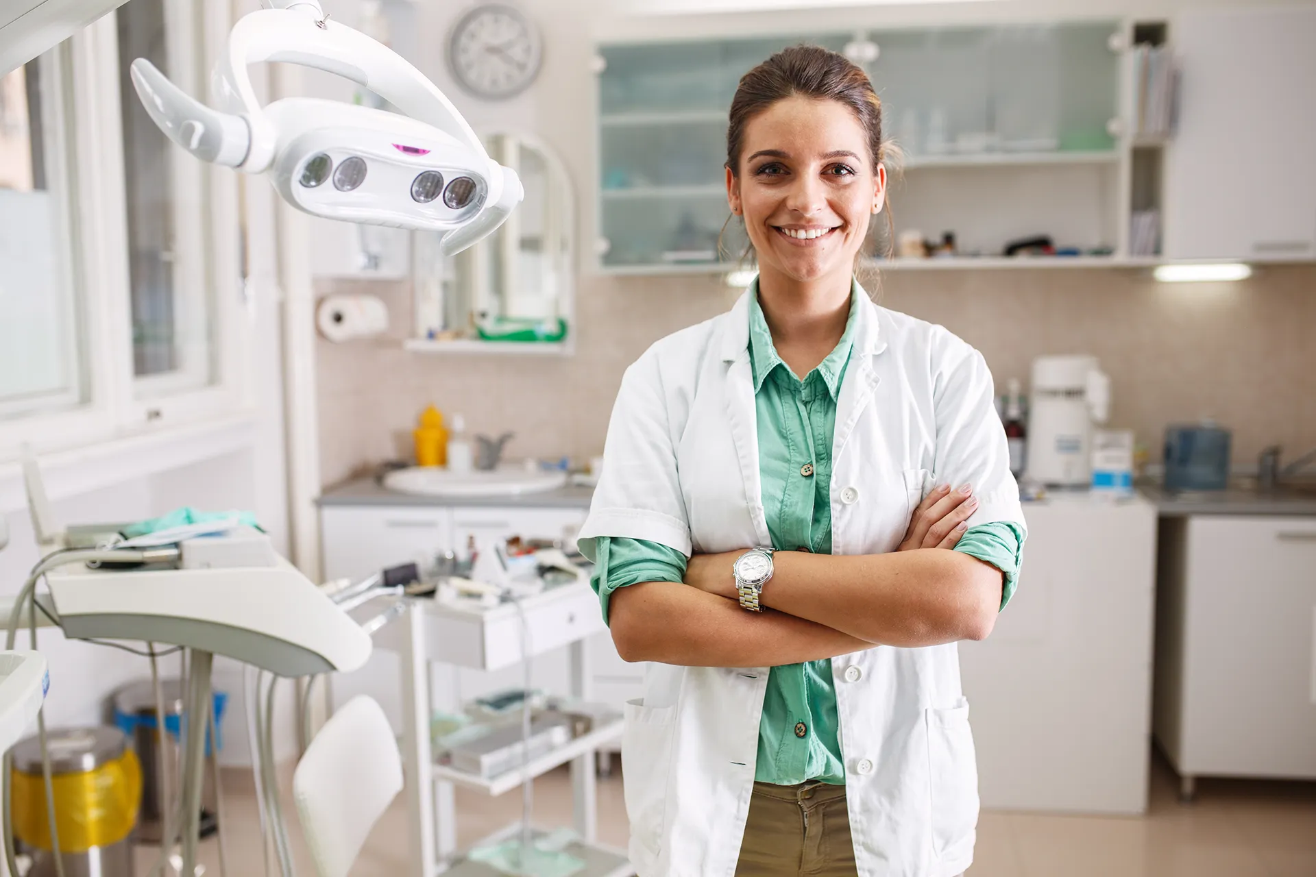 A smiling female healthcare professional in a white lab coat standing with crossed arms in a dental examination room with dental equipment and cabinetry visible
